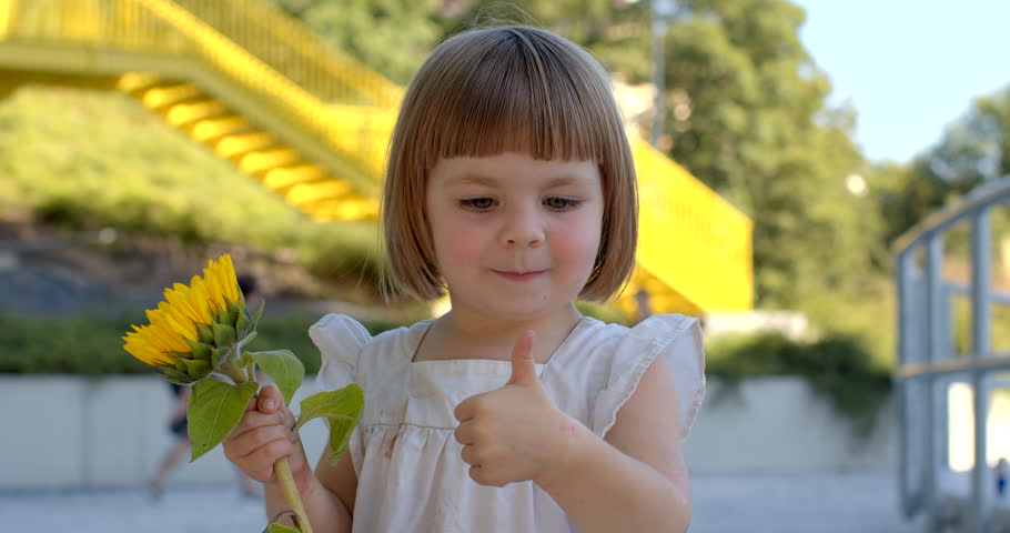 Portrait of smiling cute toddler girl with yellow sunflower, posing and giving a thumbs-up. Moments evoke a sense of joy and positivity. Hand signal used to express approval, agreement, satisfaction.