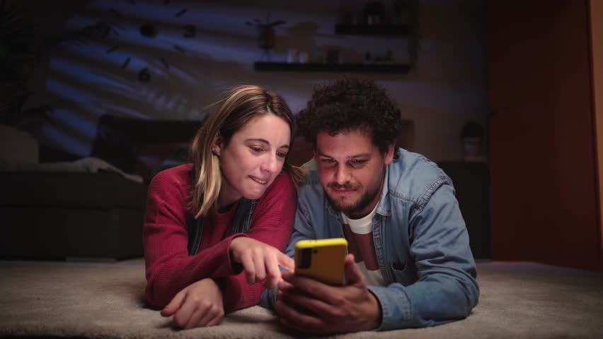 Happy Caucasian couple using animated cell phone lying on carpet in living room at home at night. Young adult couple relationship enjoying sales online shopping together. People doing domestic life. Royalty-Free Stock Footage #1109365325