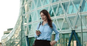 Young happy smiling business woman is using smartphone for check emails from colleagues and control agenda for meeting with partners to organise her working day while walking to the office.