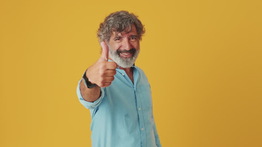 Elderly grey-haired bearded man wears a blue shirt, looks at the camera smiling and showing points with forefinger and thumbs up, isolated on orange background in studio - Powered by Shutterstock - Get 15% off with code: PIKWIZARD15
