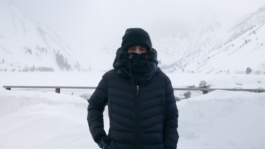 Medium shot of Indian woman in black winter clothing covering face with high fur collar during blizzard standing on the background of snowy mountains. High quality 4k footage Royalty-Free Stock Footage #1109379091