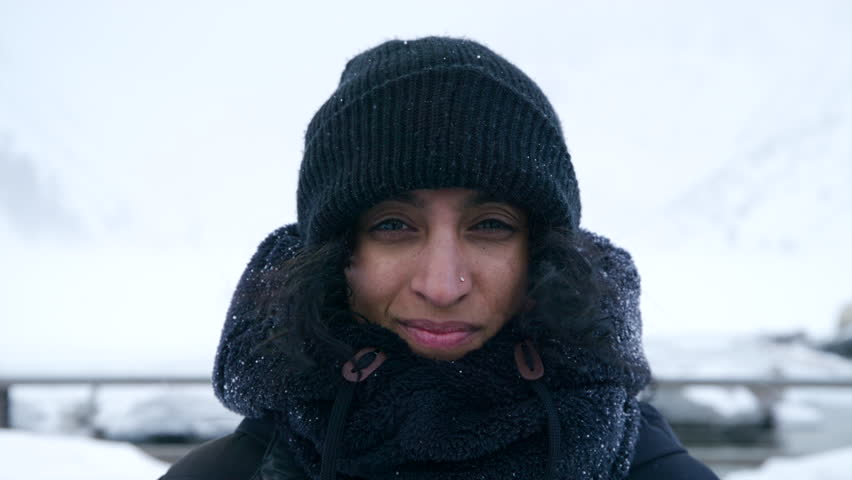 Indian pretty smiling middle-aged woman standing outdoors during snow storm. Medium close-up of Indian woman in black knitted winter hat during blizzard. High quality 4k footage Royalty-Free Stock Footage #1109379095