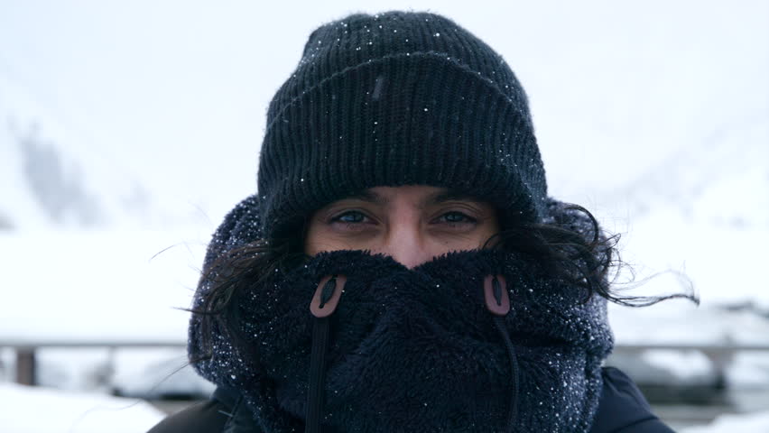Medium close-up of middle-aged Indian woman in black knitted hat, fur collar covering mouth and nose standing in mountainous area during blizzard. High quality 4k footage
