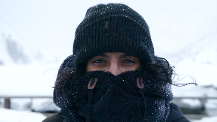 Medium close-up of middle-aged Indian woman in black knitted hat, fur collar covering mouth and nose standing in mountainous area during blizzard. High quality 4k footage Royalty-Free Stock Footage #1109379097