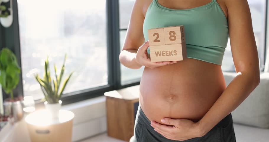 Pregnancy in early third trimester. Pregnant woman showing pregnancy belly bump for maternity photoshoot holding wooden blocks sign with 28 weeks written. Royalty-Free Stock Footage #1109388049