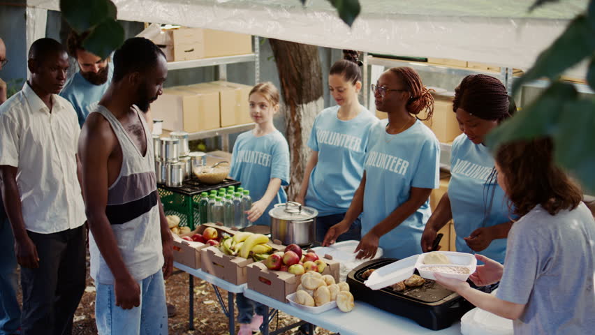 Multiethnic voluntary individuals distribute donated food, extending a helping hand to homeless and hungry people. Young volunteers share fresh, complimentary meals with those less fortunate. Royalty-Free Stock Footage #1109388145