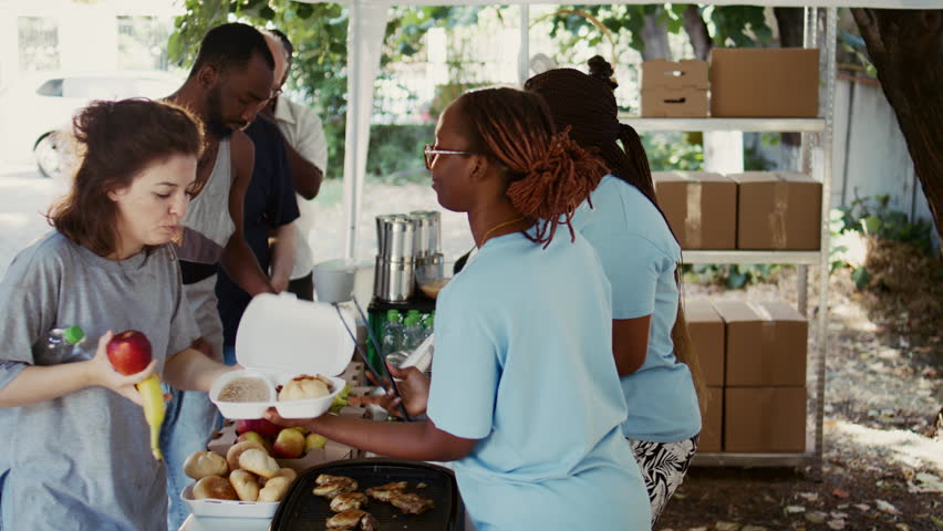 Generous volunteers wearing blue t-shirts, give free meals and humanitarian relief to those in need. Poor and homeless people of all races are fed by the non-profit organization. Side-view, handheld. Royalty-Free Stock Footage #1109388209
