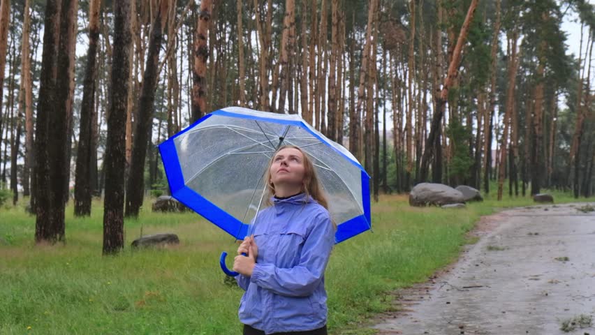 Blonde girl checking the weather under transparent umbrella outside in park. Slow motion Autumn time rainy bad forecast conditions season. Tourist with umbrella travel in new places in rain