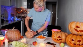 Young blonde girl stands in kitchen and cuts out pulp from pumpkin, watches video on tablet, makes her own Jack-o'-lanterns for Happy Halloween celebration at home decorated with holiday lights.