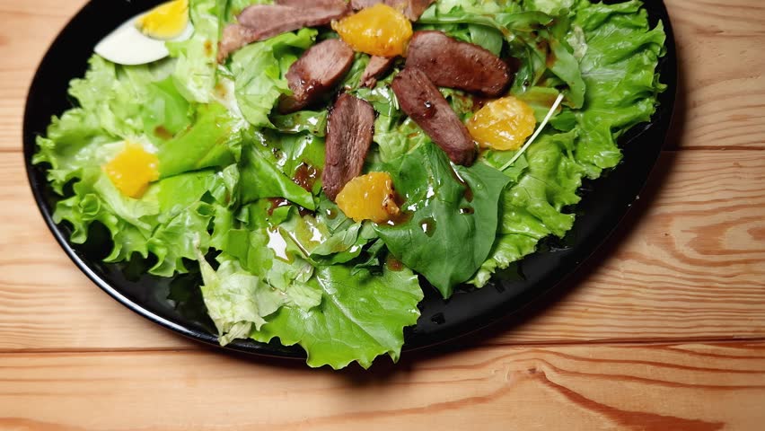 Salad made with boiled duck meat, egg, orange slices and different greens on the black dish on a rustic table, top view
