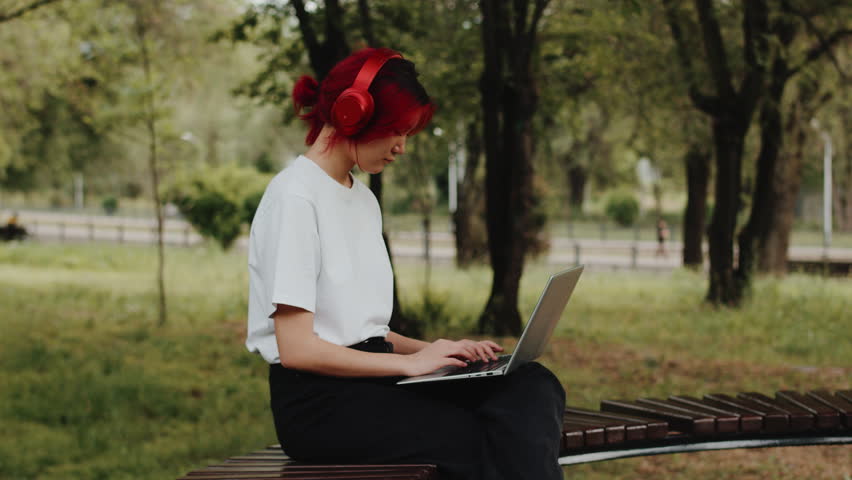 Freelancer Girl with a colourful hair sitting on a bench in a green park. Portrait of a young asian woman wearing headphones in the park during summer works using a laptop and enjoys the outdoors.