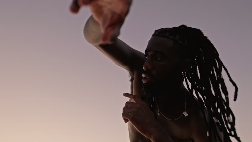 Wide low-angle shot of a shirtless black African male with dreadlocks performing conceptual dance against a sunset sky. Expressing a deep connection with his inner self Royalty-Free Stock Footage #1109407457