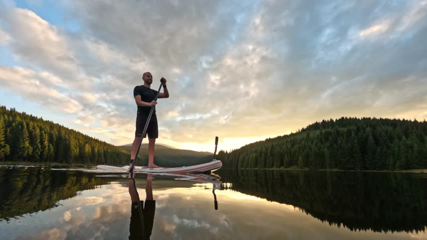 Water reflection of man silhouette standing on stand up paddle board doing sup water sport activity on a mountain lake at sunset. Healthy lifestyle outdoor activity 