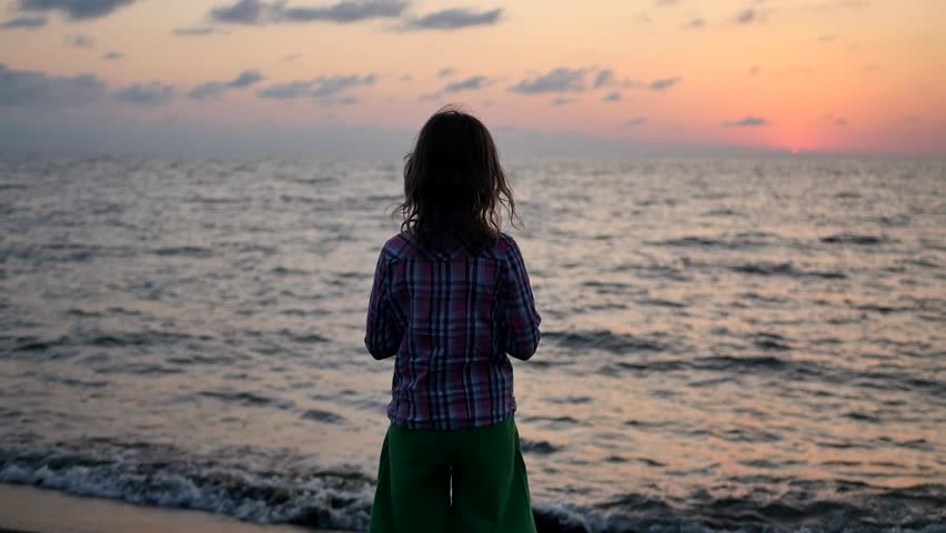Looking behind her, a little girl on the ocean shore looks at the sunset sea and admires the colorful landscape. The child looks into the distance at the horizon, waiting for the sun to rise. Royalty-Free Stock Footage #1109411687