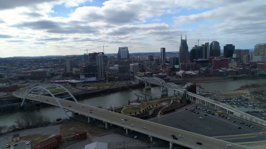 Aerial Panning Shot Of Vehicles On Bridge Above River Near Buildings In City Against Cloudy Sky - Nashville, Tennessee