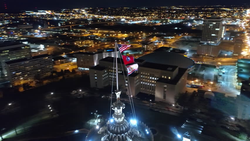 Aerial Panning Shot Of Flags On Tennessee State Capitol Spire Amidst Buildings In City At Night - Nashville, Tennessee