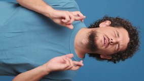 Vertical video, Guy with curly hair dressed in blue t-shirt holds his fingers crossed and makes wish, isolated on blue background in the studio
