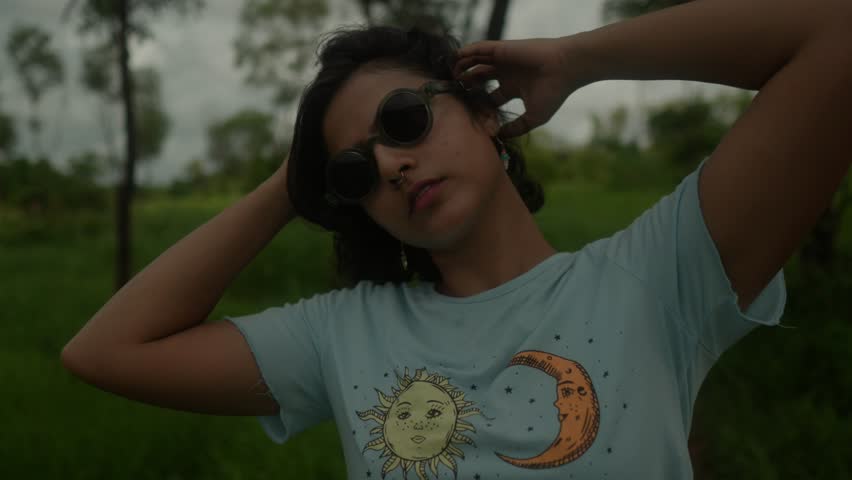 Close-up shot of an Indian young girl surrounded by the forest of India, she takes a moment to comb her hair while wearing stylish sunglasses. With a graceful turn she looks directly into the camera.