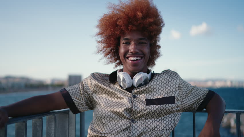 Portrait of Brazilian young man with stylish curly hair, smiling sincerely at camera while recreating at seashore. Youth trendy African American guy with piercing, feeling happy, calm and carefree