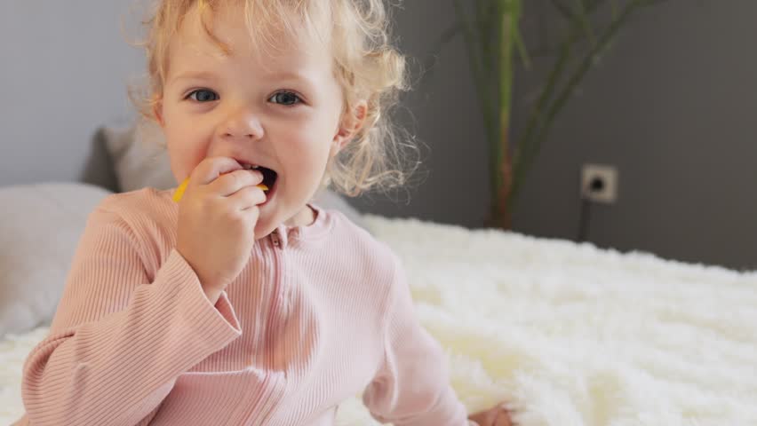 Toddler wavy haired blond girl sitting in bed eating dried apricots with pleasure wearing ping attire enjoying delicious snack expressing positive emotions.