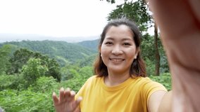 Positive female hipster waving a hand at the camera during video chat online. Smiling hiker gesturing hands. Cheerful traveler exploring nature.