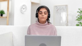 Modern Muslim woman in headphones talking online video call use laptop cyberspace chatting at white room. Brunette female internet remotely working consulting communication videoconference webinar