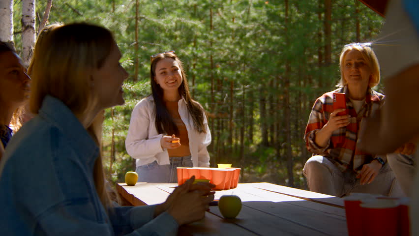 Young people play beer pong. Stock footage. Game for group of friends with glasses of alcohol and ping pong ball. Friends play beer pong in nature in summer