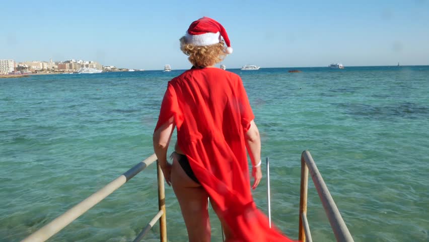 middle-aged  woman in santa claus hats and sunglasses wearing in swimsuit on the sea beach, women dancing and wishes a merry Christmas and happy new year