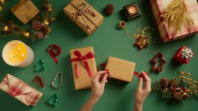 Overhead view of woman tying red silk ribbon on golden gift box. Top down shot female packing present at green background. Overhead flat lay of Christmas art and craft equipment surrounding lady hands