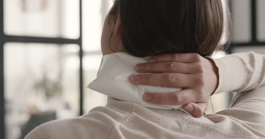 back view of a suffering woman trying to relieve neck pain with an ice pack at home.