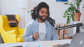 Young cheerful Arabian man journalist runs own audio podcast on internet talking about news and actively gesturing with cup of coffee in hands sits in home office at table with microphone and laptop