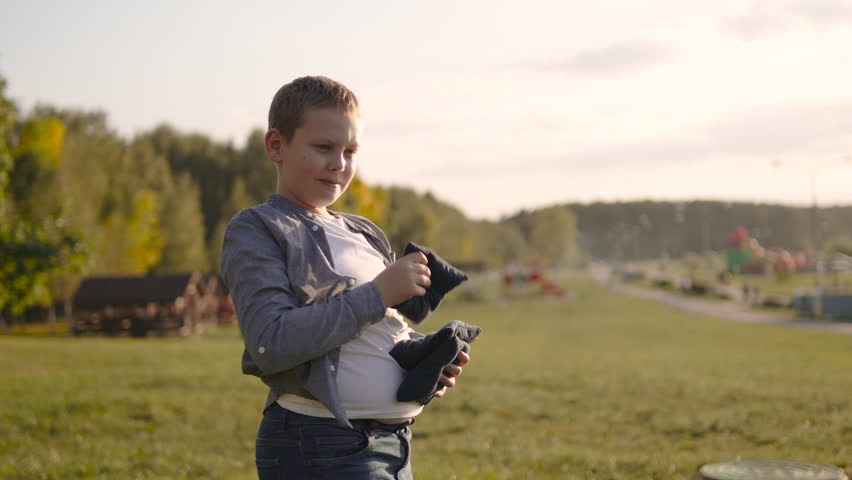 Boy plays cornhole, throwing bean bags and reacting emotionally to misses and hits. Game of accuracy competition in sunny weather on lawn. Portrait of diligent overweight child having fun in sunlight.