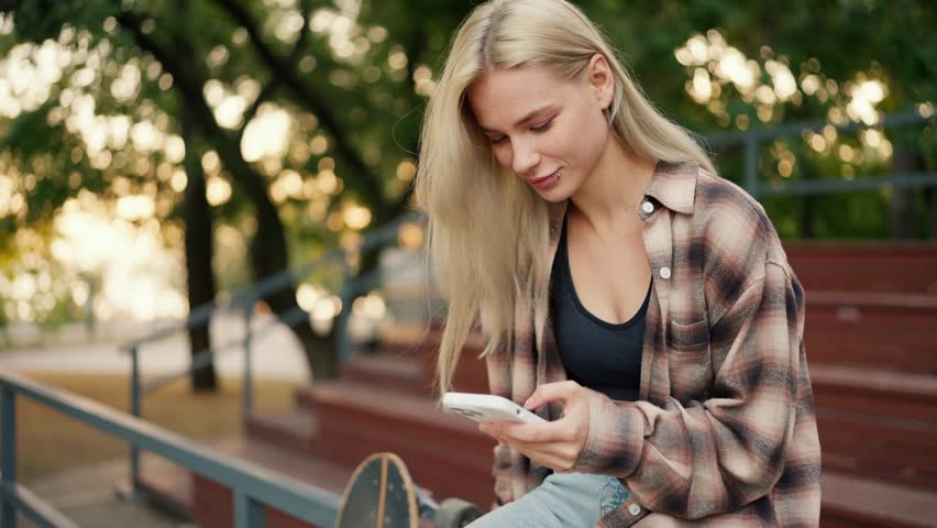 A blonde girl in a plaid shirt and black top is typing something on her white phone while on the brown stands at a skatepark in the summer. Happy blonde teenager girl watching something on her white Royalty-Free Stock Footage #1109447549