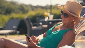 Senior retired woman wearing swimming costume, sun hat and sunglasses relaxing by hotel pool on lounger looking at social media on mobile phone on summer holiday - shot in slow motion - Powered by Shutterstock - Get 15% off with code: PIKWIZARD15