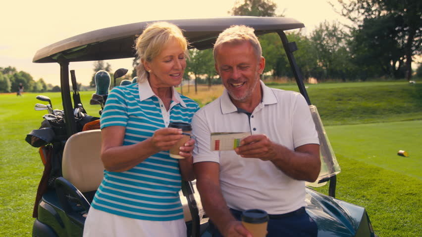 Senior retired couple next to buggy having takeaway coffee during round of golf looking at score card together - shot in slow motion Royalty-Free Stock Footage #1109448225