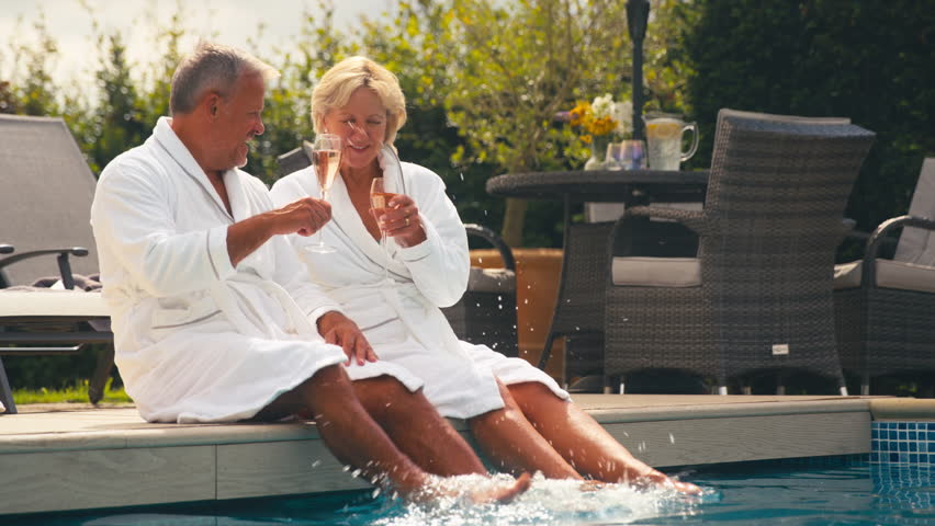 Senior retired couple wearing robes drinking champagne and splashing in hotel swimming pool on summer spa holiday - shot in slow motion Royalty-Free Stock Footage #1109448301