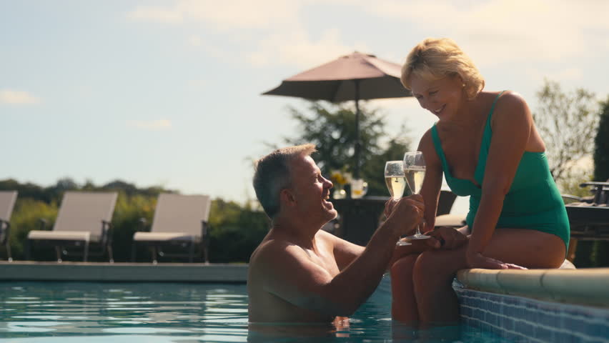 Senior retired couple on holiday with man in water and woman sitting on edge of hotel pool making toast with champagne together - shot in slow motion Royalty-Free Stock Footage #1109448311