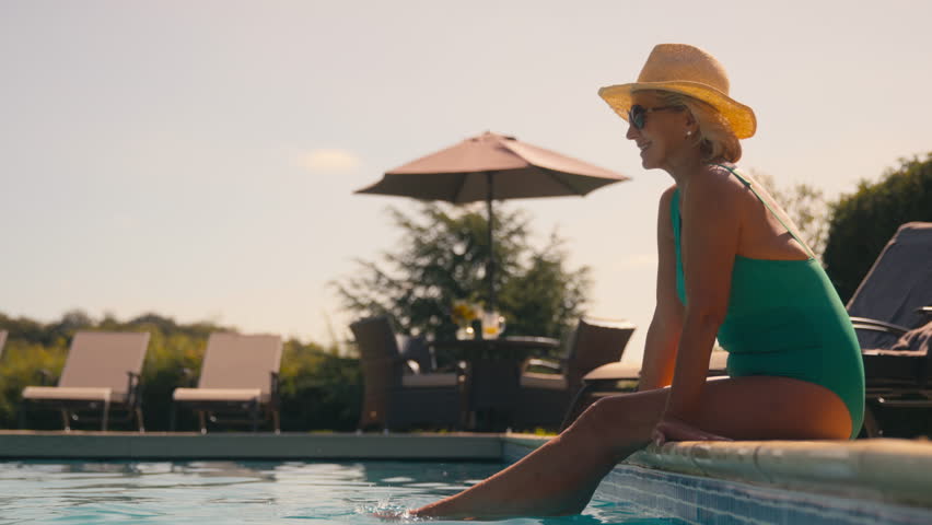 Senior retired couple on holiday with man swimming to woman sitting on edge of hotel pool - shot in slow motion Royalty-Free Stock Footage #1109448315