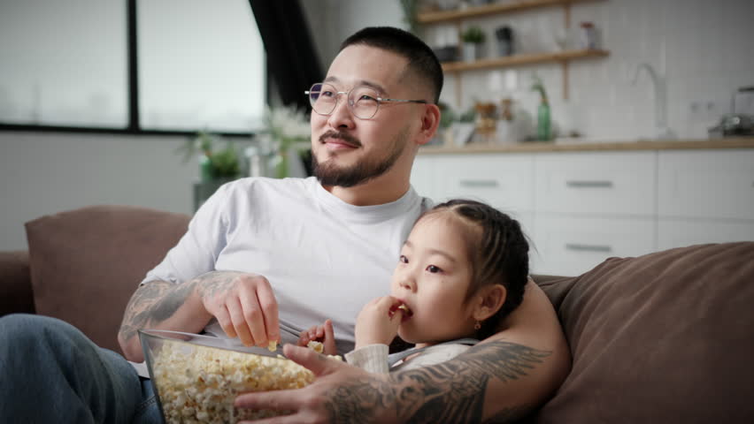 Dad And Daughter Watching TV Eating Popcorn. Korean family having fun sitting on couch at home. Loving father hugging little daughter. 