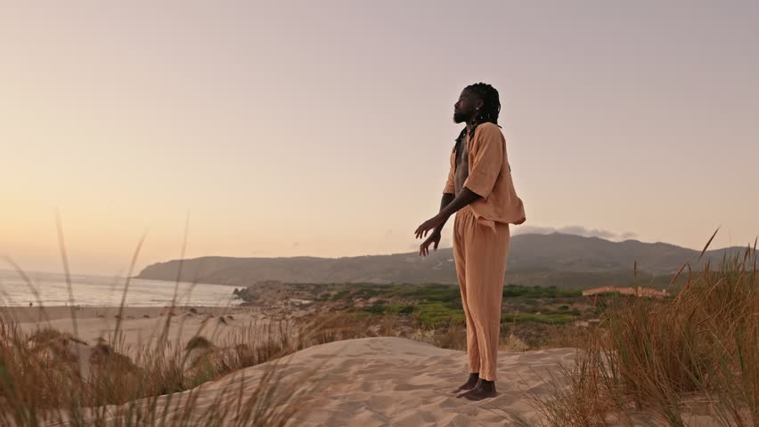 An artistic black man in loose attire stretches his hands at sandy beach dunes during a beautiful sunset, connecting deeply with the motherland Royalty-Free Stock Footage #1109459577