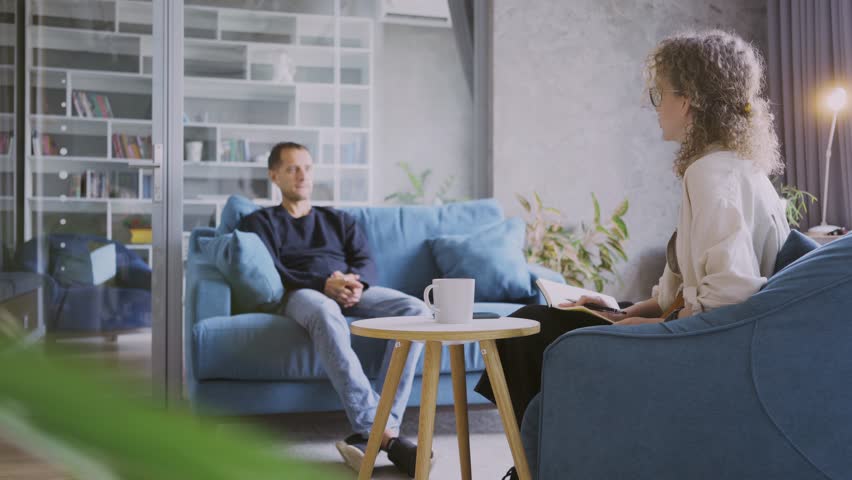 Back side view woman with open paper notebook and drink mug sits opposite and listens to man on comfortable sofa with cushions at an appointment in the company office  Royalty-Free Stock Footage #1109470379