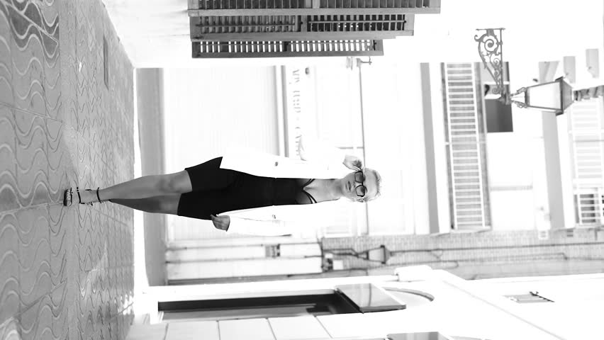 Fashionable blonde with short hair and sunglasses in a white jacket on the street on a sunny day. Black and white vertical video.
