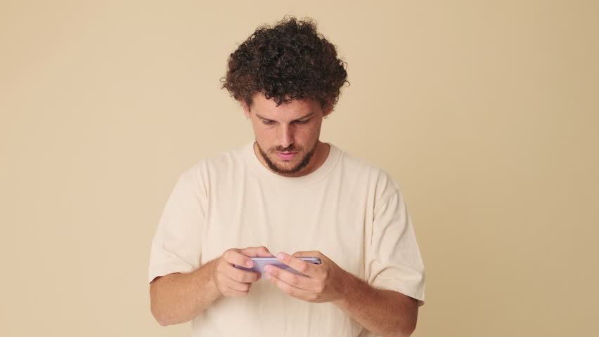 Guy with curly hair dressed in beige T-shirt, playing online video game, wins, waves hands triumphantly gesture standing in studio on beige background in the studio