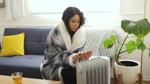 Latin woman with curly hair wrapped up in a blanket and sitting next to a radiator rubbing her hands to stay warm during winter and cold weather at home Adlı Stok Video