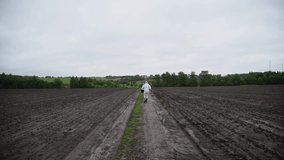 Woman in white clothing with bucket walks on rural dirt road between agricultural plowed field (after wheat harvesting) in a cloudy morning. Overcast sky. Real time video. Farming theme.