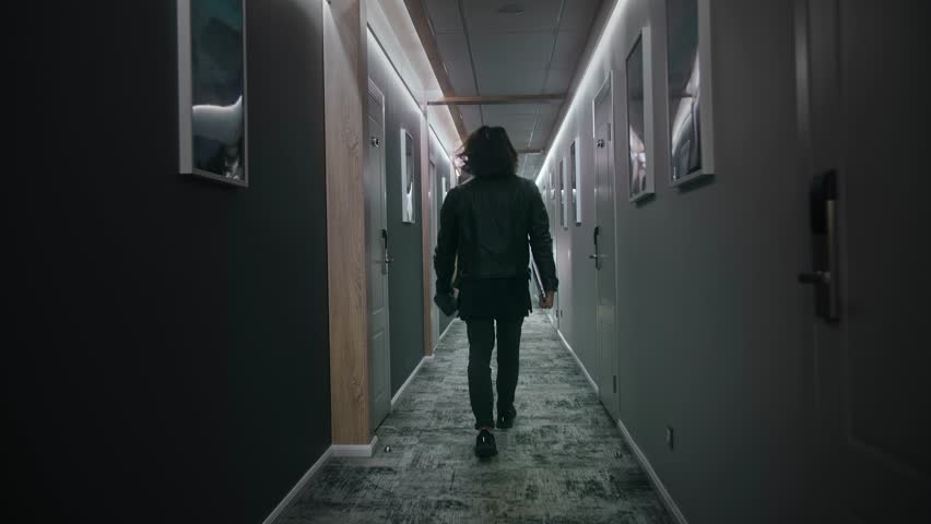 A long-haired man is walking down the corridor of a hotel floor. He is wearing dark jeans and a leather jacket, holding a camera and a laptop. Dim light, camera in motion