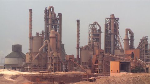 A pan of a working cement factory in an industrial area in Salalah in Oman, showing moving trucks and a port in the distance.