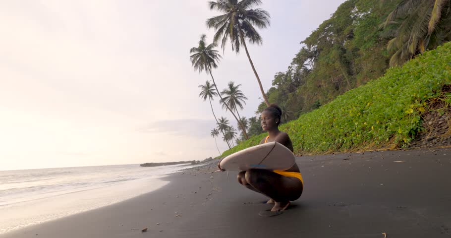 Gorgeous young black girl happily sitting on the beach with her surfboard in Sao Tome and Principe, Africa