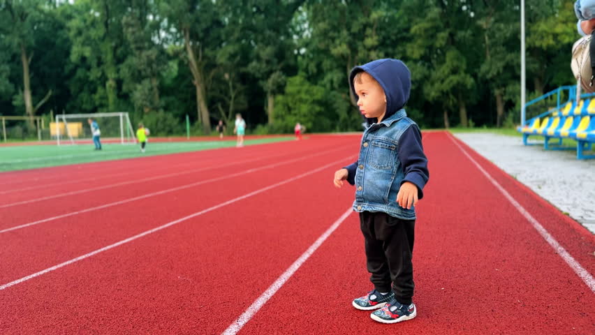 Focused Caucasian boy stands at the stadium. Kid watches the children playing sports games.
