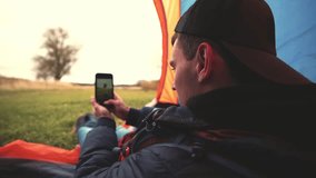Young guy sits in tent in nature and shoots video on smart phone or takes photo of outdoors landscape with river or lake using camera. Male breaks camp on hike or picnic, sits under an awning alone.